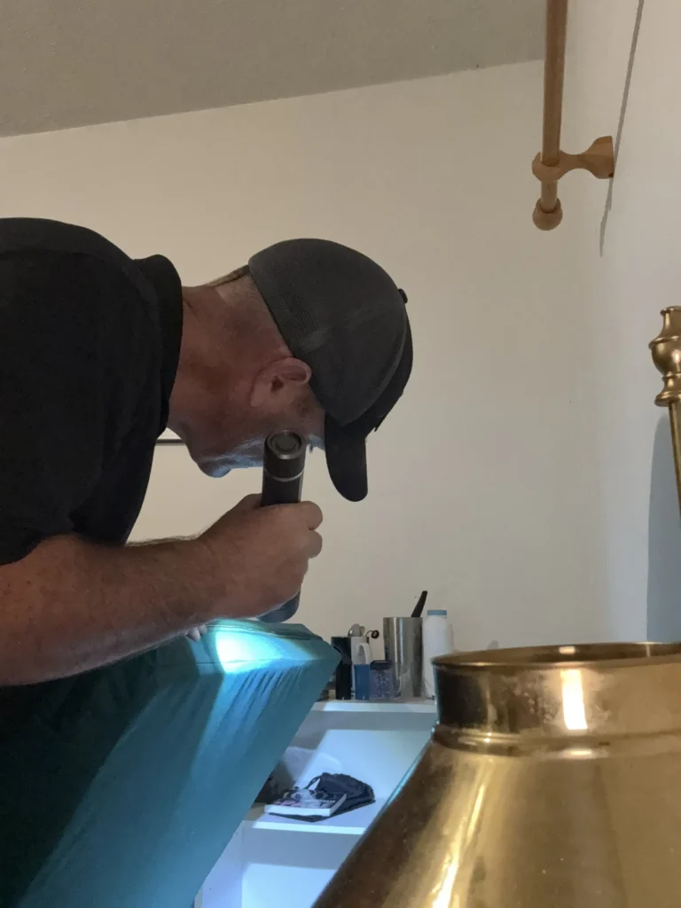 This is a close-up photograph of a man in what appears to be a bedroom, seemingly conducting a bed bug inspection.