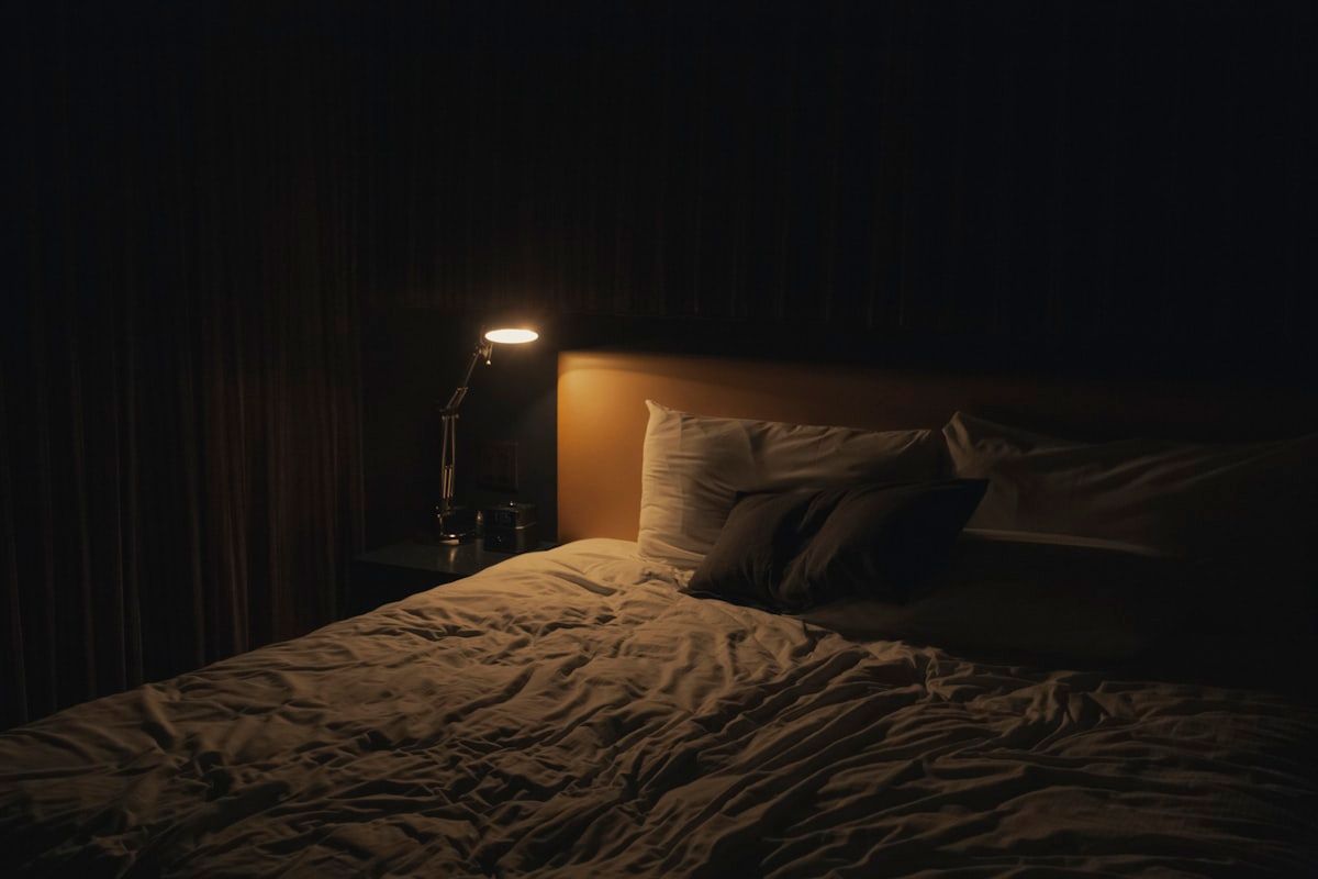 A dimly lit bedroom with a flashlight shining along mattress seams while a hand uses a stiff card to lift fabric folds, with a sealed zip-top bag and a small jar placed on the bed for collecting a sample