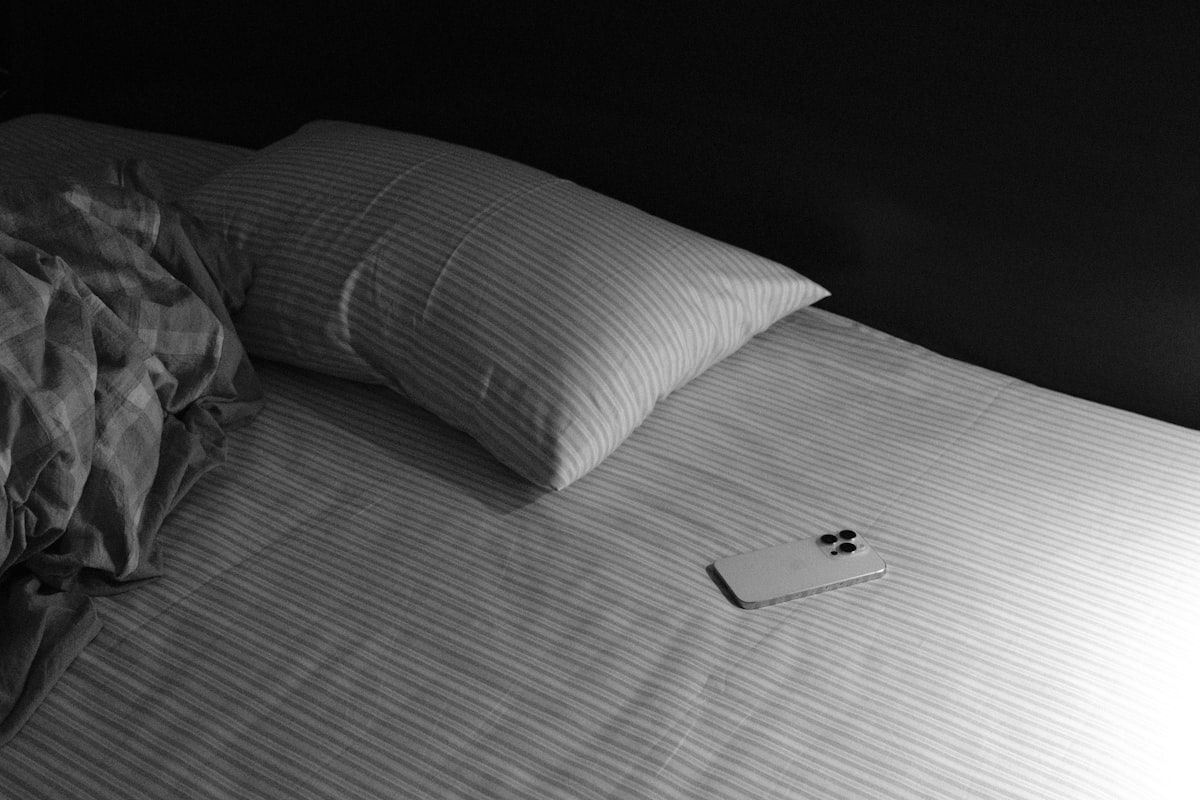 A careful home inspection setup on a bed with a flashlight, a credit card-sized plastic card used to probe mattress seams, a small sealed container holding a specimen, and a phone camera positioned close to document a bug found in the fabric folds