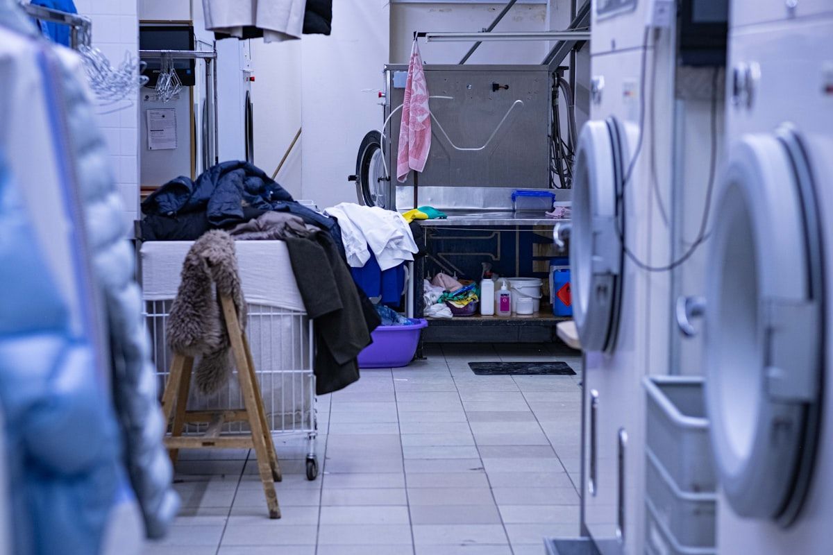 A laundry area with sheets and pillowcases being placed into sealed plastic bags next to a washer and dryer, while a bed in the background remains untouched and neatly made to show containment before cleaning