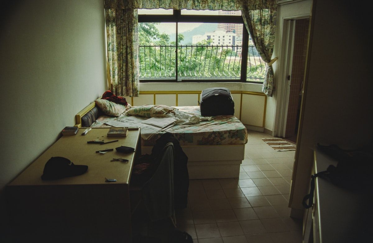 A messy bedroom with an unmade bed, cluttered floor, an open closet packed with clothes, a sofa pushed against the wall, and a technician inspecting baseboards and furniture seams with a flashlight