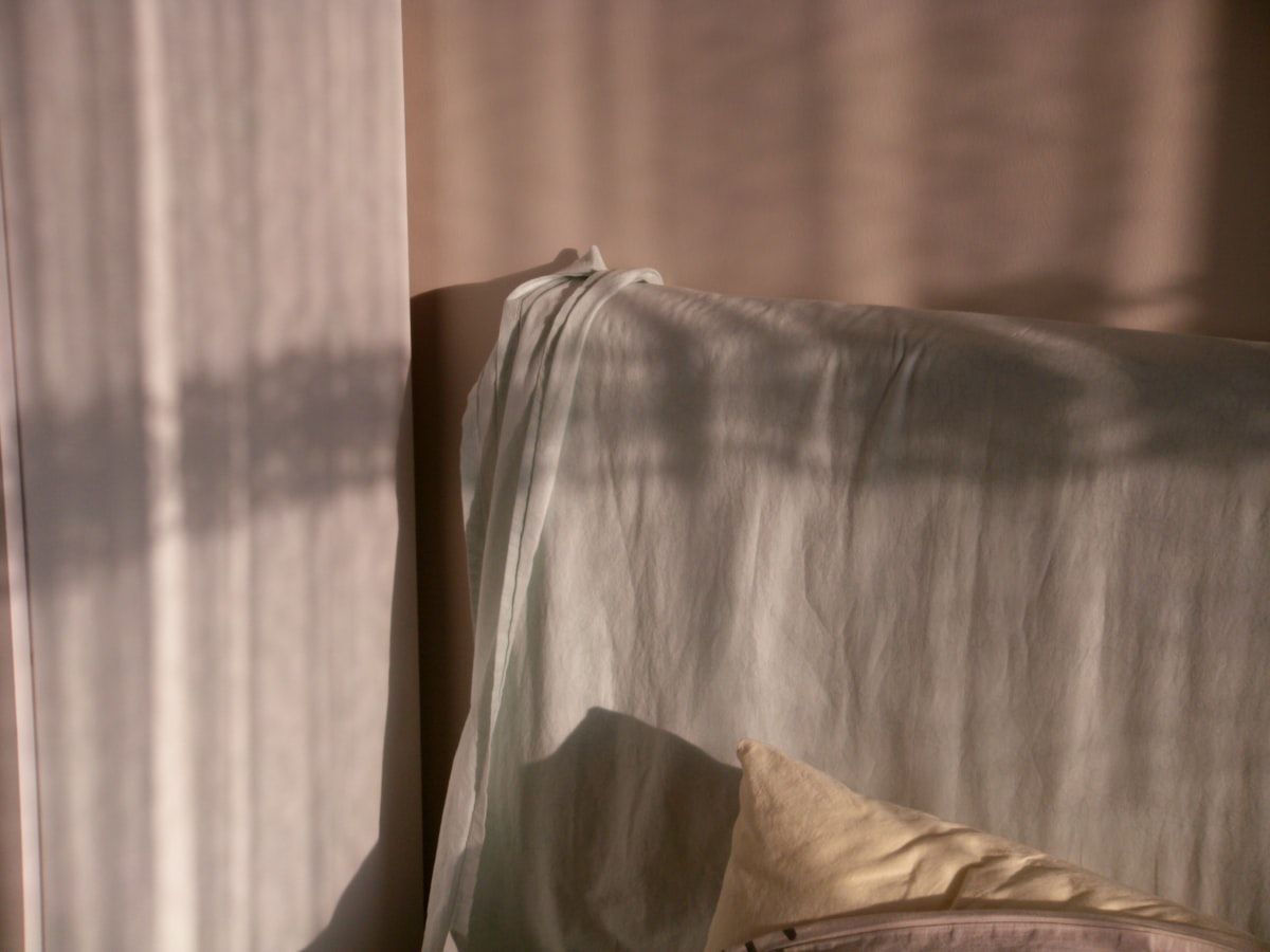 A bed frame, headboard, and nearby nightstand pushed close together against a bedroom wall, with a flashlight revealing tiny pale eggs in the mattress piping, screw holes in the wooden frame, and cracks along the baseboard.